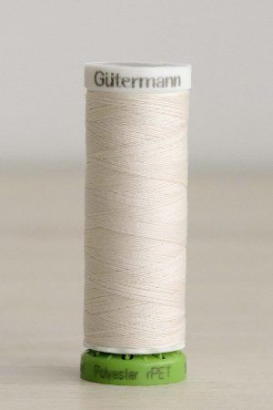 Bobina de hilo Gütermann 135 rPET, de blanco crema, sobre una madera de color claro y de fondo una pared blanca.