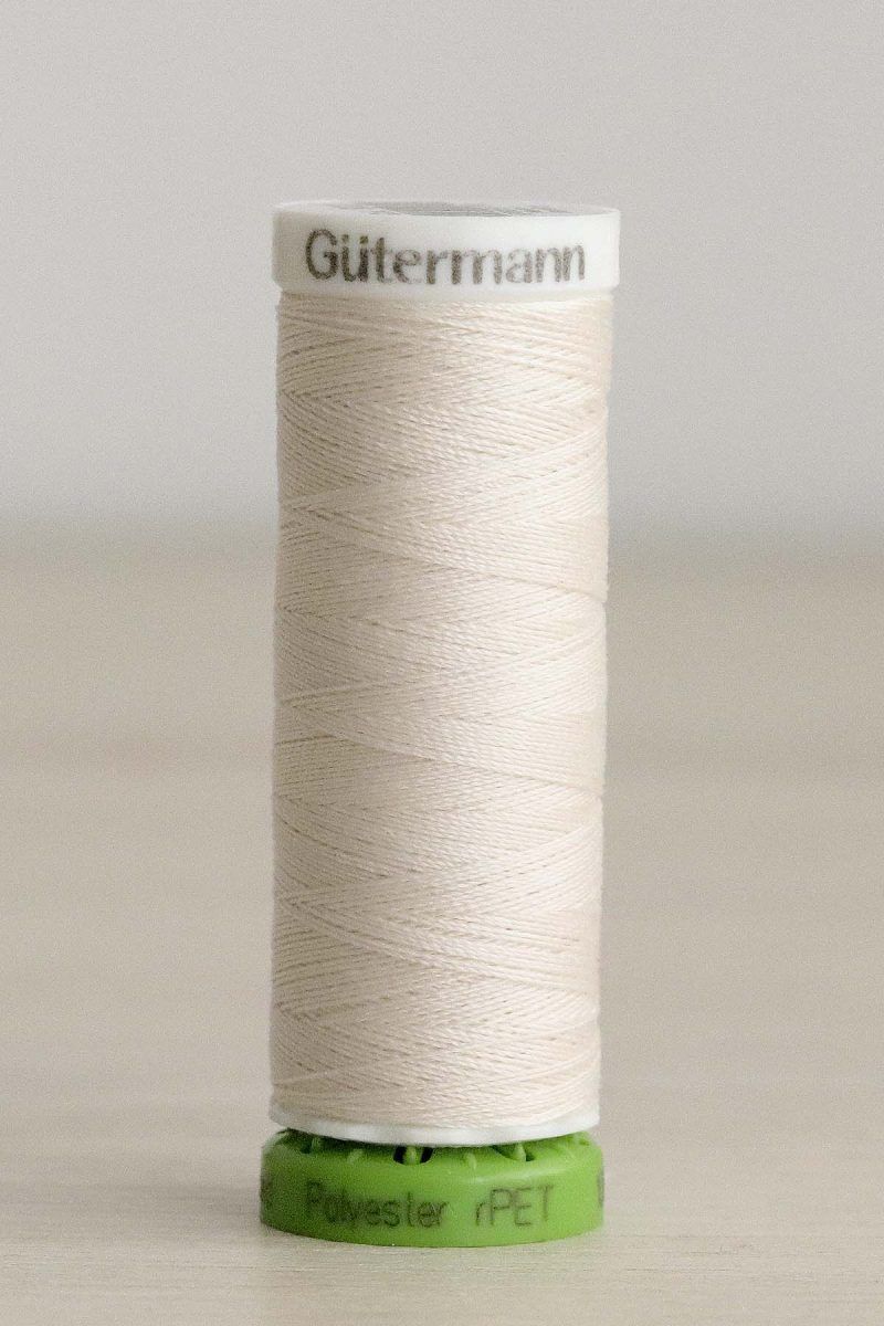 Bobina de hilo Gütermann 135 rPET, de blanco crema, sobre una madera de color claro y de fondo una pared blanca.