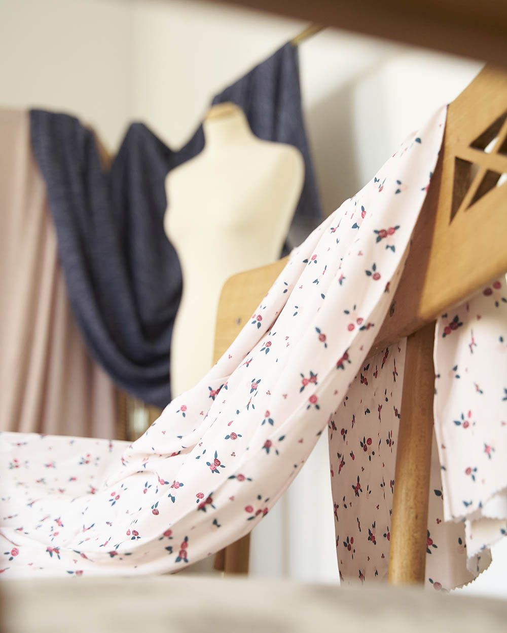 Una tela de color blanco crema, con estampado de ramilletes de flores pequeñas en tonos rojos y rosas con hojas verdes, se encuentra sobre un mantel de esparto. En el fondo del plano vemos un maniquí para el diseño de textiles y costura de alta calidad, así como muestras de la misma colección de telas en colores más oscuros.