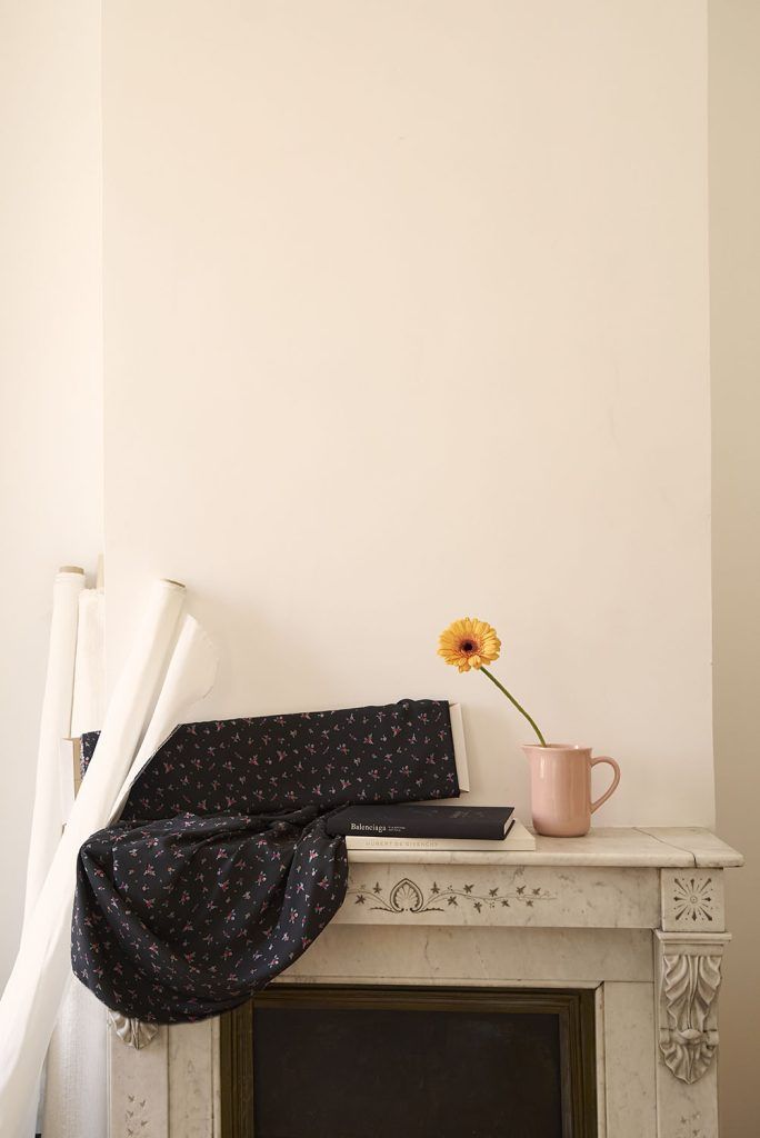 Una tela de color negro, con estampado de ramilletes de flores pequeñas en tonos rojos y rosas con hojas verdes pastel sobre la encimera de la chimenea. En el primer plano aparece una flor amarilla que contrasta con el delicado estampado de la tela de muestra.