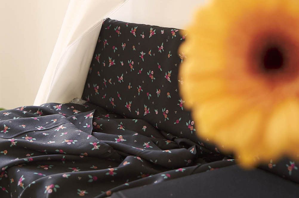 Una tela de color negro, con estampado de ramilletes de flores pequeñas en tonos rojos y rosas con hojas verdes pastel. En el primer plano aparece una flor amarilla ligeramente desenfocada.