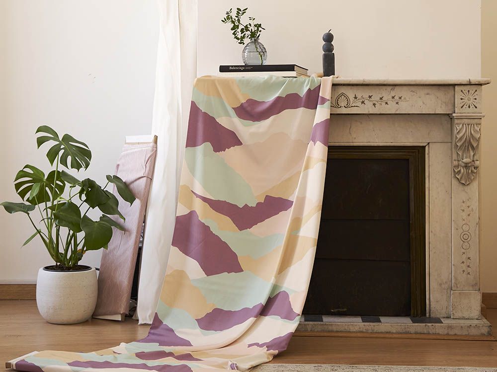 Sobre una chimenea de mármol cuelga una tela con estampado de montañas en colores morado, beige, rosa y verde azulado. A su lado hay un rollo de tela blanco, una tablilla con tela rosa y una maceta con una planta.