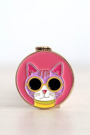 Porta agujas imantado. Tiene forma de bastidor de bordar redondo. Dentro de esa bastidor, sobre un fondo rosa, está la cara de un gatito rosa clarito con gafas de sol con montura amarilla.