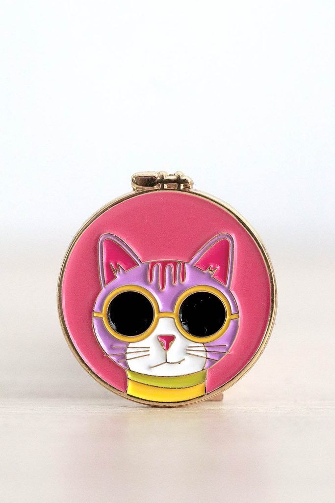 Porta agujas imantado. Tiene forma de bastidor de bordar redondo. Dentro de esa bastidor, sobre un fondo rosa, está la cara de un gatito rosa clarito con gafas de sol con montura amarilla.