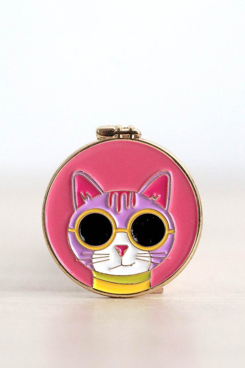 Porta agujas imantado. Tiene forma de bastidor de bordar redondo. Dentro de esa bastidor, sobre un fondo rosa, está la cara de un gatito rosa clarito con gafas de sol con montura amarilla.