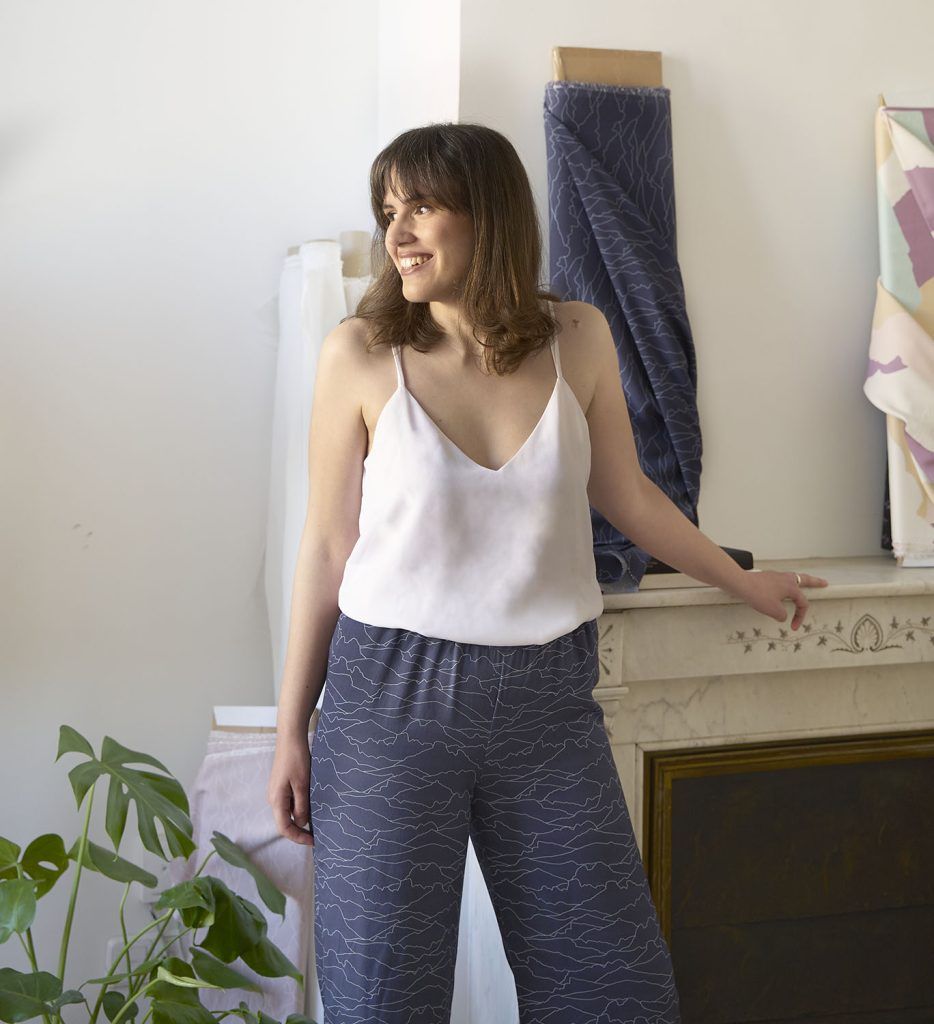 Mujer con amplia sonrisa y pelo castaño modela de perfil un diseño de pantalón con tela sostenible de color azul y patrones de líneas blancas que emulan el horizonte.