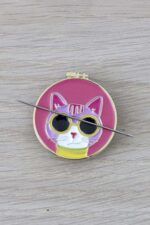 Porta agujas gato con aguja Porta agujas de color rosa con forma de bastidor de bordar. En él aparece la cara de un gato rosa claro, con gafas de sol y camiseta amarillas. Sobre el porta agujas se posa una aguja de coser a mano.