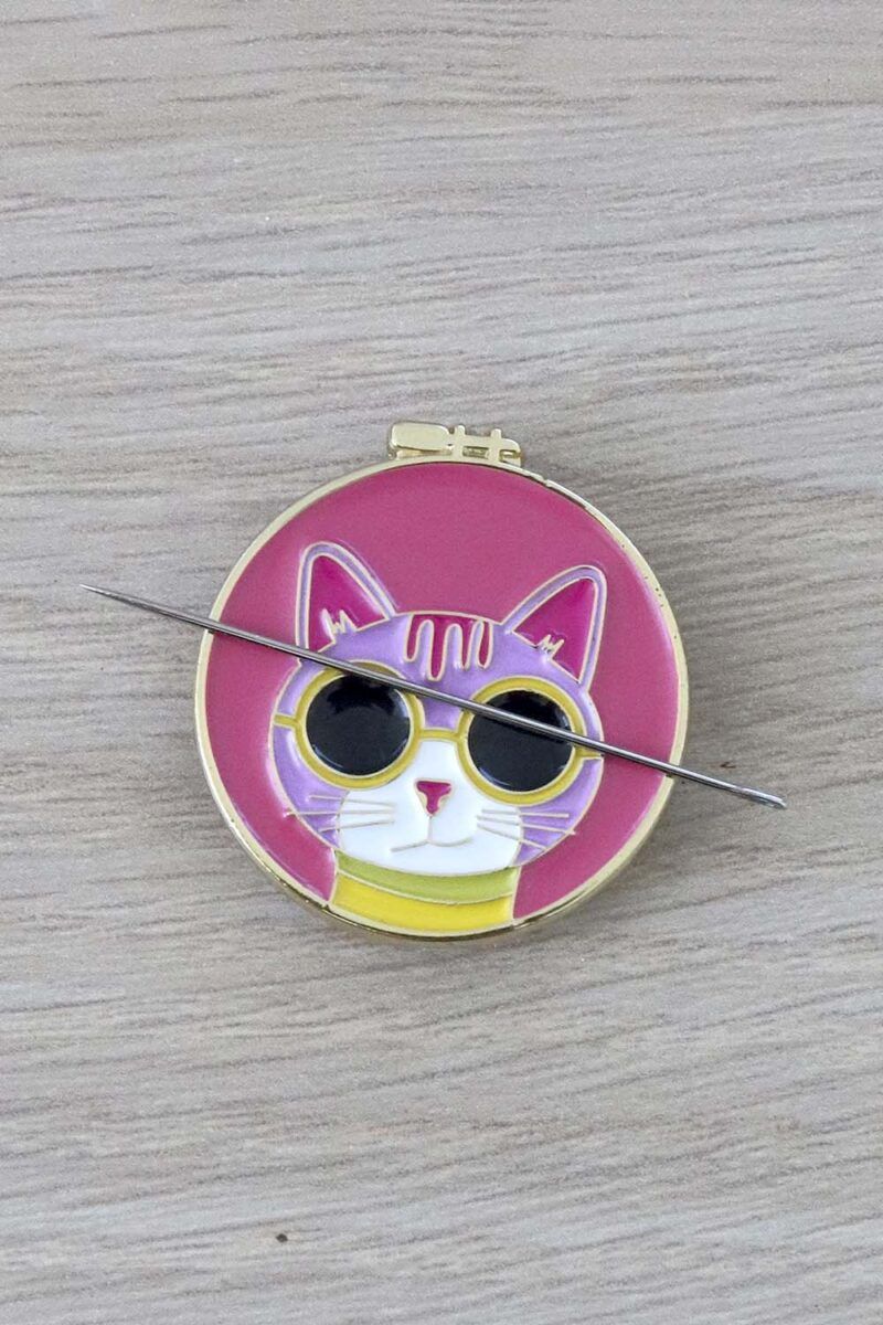 Porta agujas de color rosa con forma de bastidor de bordar. En él aparece la cara de un gato rosa claro, con gafas de sol y camiseta amarillas. Sobre el porta agujas se posa una aguja de coser a mano.