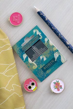 Tres porta agujas imantados con forma de bastidor redondo están encima de una mesa de madera. Uno tiene una cara de gatito, otro una ramo de flores y en otro se lee "son of a stitch". A su lado se encuentra un pack de agujas con packing de caballitos de mar y corales, un lápiz de tiza para marcar la tela con motivos de constelaciones y una tela color mostaza con líneas blancas que forman montañas.