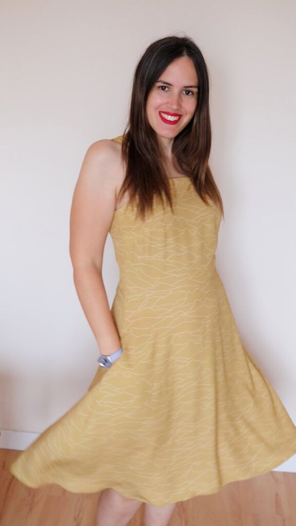 Mujer castaña de pelo largo posa sonriente frente a la cámara con un vestido mostaza de tirante con pequeñas líneas blancas que conforman las siluetas de diferentes montañas.