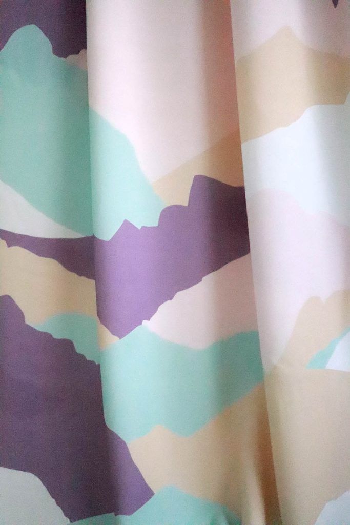 Plano cercano de una tela de viscosa que cae haciendo varios pliegues. Tiene un estampado de siluetas de montañas en colores morado, crema, marrón claro, verde agua, blanco y rosa pálido.