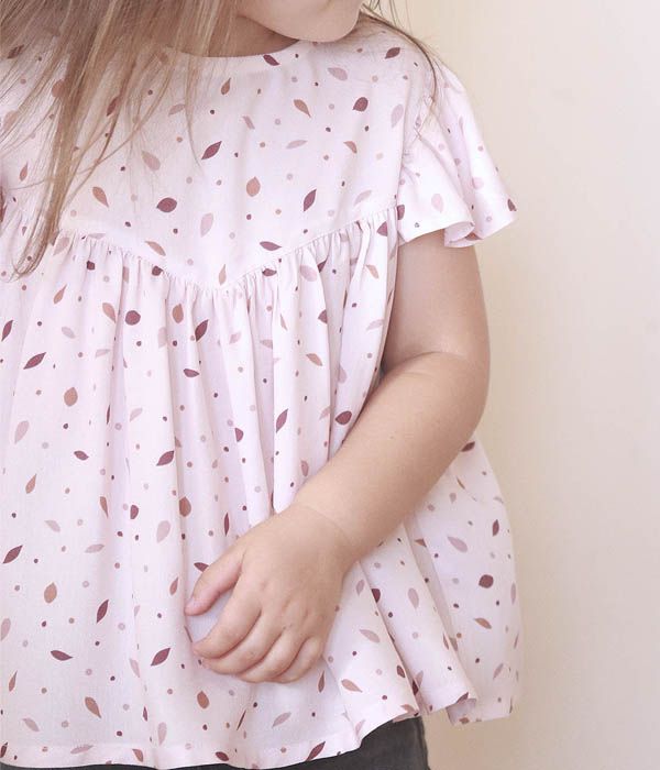 Una niña pequeña luce un hermoso vestido de delicados pliegues con tela sostenible de color rosa claro y patrones de hojas que evocan una suave brisa.