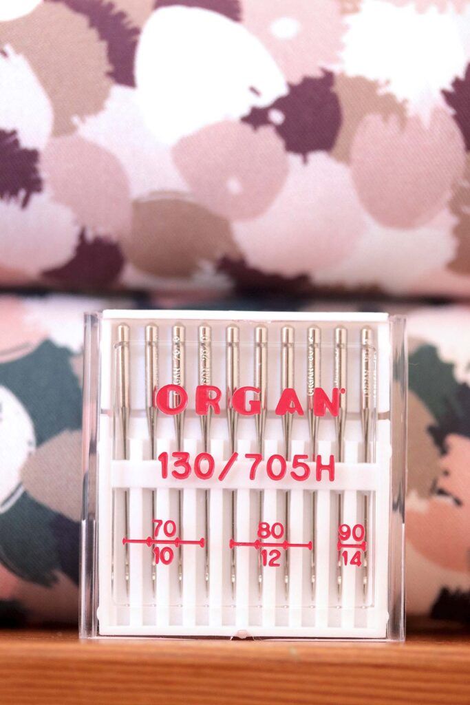 Cajita blanca con surtido de 100 agujas de coser a máquina universales. En la caja se puede leer en rojo la marca, ORGAN, y los grosores de la aguja: 70, 80 y 90. Al fondo de ven dos tablillas de telas. Una de flores rosas, marrones, blancas y burgundy y otra con el mismo estampado, pero en colores verdes, rosas y blancos
