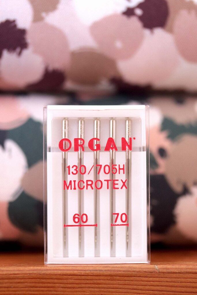 Cajita blanca con surtido de 5 agujas de coser a máquina. En la caja se puede leer en rojo la marca, ORGAN, el tipo de aguja (Microtex) y los grosores de la aguja: 60 y 70. Al fondo de ven dos tablillas de telas. Una de flores rosas, marrones, blancas y burgundy y otra con el mismo estampado, pero en colores verdes, rosas y blancos.