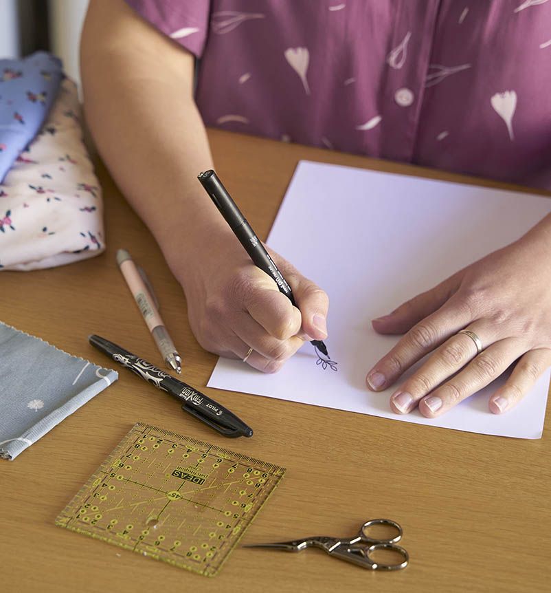 Plano cercano en el que se ven las manos de Lucía dibujando con rotulador negro una pequeña rama sobre un folio blanco. En la mesa, de color marrón, aparecen una tela azul y otra blanca, ambas con flores. También hay una tijera, una regla cuadrada y un bolígrafo.