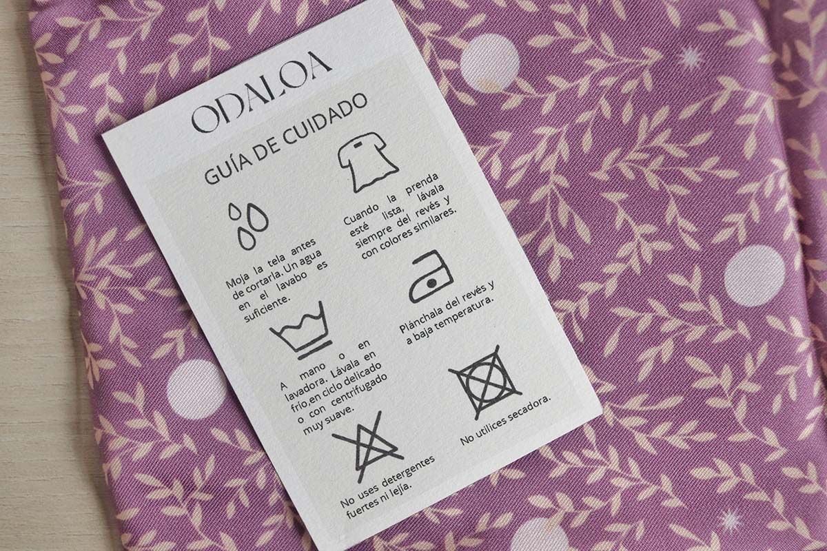 Sobre una tela doblada, de color morado, con hojitas color arena y topitos blancos, se encuentra una etiqueta de la marca ODALOA en la que se identifican los símbolos de cuidado de la tela. El primero de ellos son unas gotitas de agua e indica que hay que lavar la tela antes de usarla. Luego aparece un símbolo de una camiseta del revés y señala que hay que lavar las prendas de esta manera. A continuación aparece un símbolo de lavado en frío y delicado y aclara que hay que lavarla así. Después aparece el icono de una plancha con un punto en su interior y describe que hay que planchar la prenda a baja temperatura y del revés. El penúltimo símbolo es un triángulo tachado con una X, que indica no utilizar lejía ni blanqueantes. Por último, un símbolo conformado por un cuadrado, dentro de éste un círculo y todo ello tachado con una x. Y se indica que no hay que utilizar secadora para secar ese tejido.