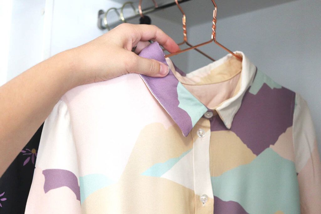 Una mano de mujer sujeta una percha metálica de color cobre sobre la que cuelga una camisa de mujer con un estampado abstracto de montañas en colores morado, verde y crema pastel. Está colgando la blusa en un armario cuyo interior es de color blanco.