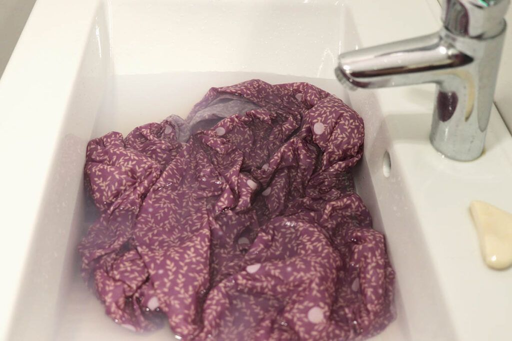 En un lavabo rectangular de color blanco se encuentra a remojo una tela morada con un estampado de hojitas color arena y topitos blancos. Sobre el lavabo se encuentra una pastilla de jabón.