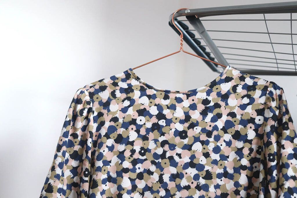 Plano cercano de una blusa con estampado de flores abstractas en colores azules, rosas, blancos y ocres está tendida en un tendero de interior metálico. La percha que sujeta blusa es metálica y de color cobre. El tendedero se encuentra dentro de una habitación con paredes blancas.