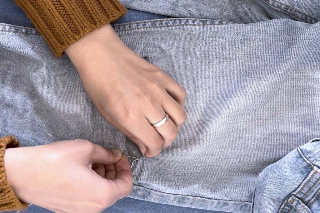 La imagen ilustra el remiendo de un pantalón vaquero en la zona de la rodilla. Unas manos de mujer sujeta una aguja mientras la clava en la tela vaquera, la otra mano sujeta el pantalón. Está cosiendo, de manera que quede visible, un parche para tapar un agujero.