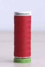 Bobina de hilo Gütermann 156 rPET, de color rojo intenso, sobre una madera de color claro y de fondo una pared blanca. La bobina es de plástico verde brillante en la parte inferior y blanca en la parte superior, y en donde se puede leer "Gütermann".