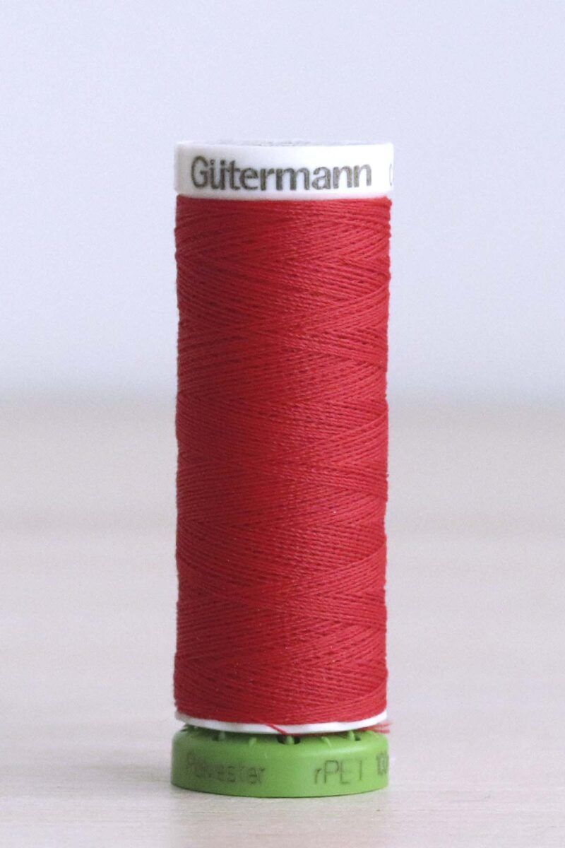 156_Hilo_rPET_Gütermann_poliéster_Huellas_rojo Bobina de hilo Gütermann 156 rPET, de color rojo intenso, sobre una madera de color claro y de fondo una pared blanca. La bobina es de plástico verde brillante en la parte inferior y blanca en la parte superior, y en donde se puede leer "Gütermann".