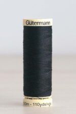 Bobina de hilo Gütermann 665, de color azul marino casi negro, sobre una madera de color claro y de fondo una pared blanca. En la parte superior de la bobina, de color crema, se puede leer "Gütermann".