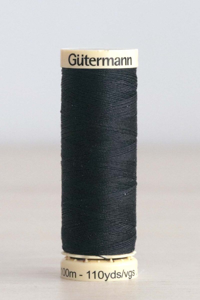 665 Hilo Gütermann poliéster Guijarros Bobina de hilo Gütermann 665, de color azul marino casi negro, sobre una madera de color claro y de fondo una pared blanca. En la parte superior de la bobina, de color crema, se puede leer "Gütermann".