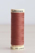 Bobina de hilo Gütermann 847, de color marrón arcilla, sobre una madera de color claro y de fondo una pared blanca. En la parte superior de la bobina, de color crema, se puede leer "Gütermann".