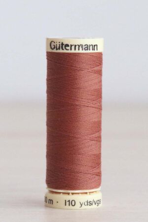Bobina de hilo Gütermann 847, de color marrón arcilla, sobre una madera de color claro y de fondo una pared blanca. En la parte superior de la bobina, de color crema, se puede leer "Gütermann".