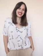 Una chica de pelo largo, castaño, posa sonriente frente a la cámara. Lleva una blusa abotonada, de manga corta, blanca y con estampado de caras abstractas en color azul oscuro. Lleva las manos dentro de los bolsillos del pantalón, de color beige.