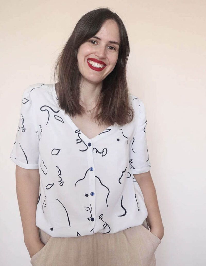 Una chica de pelo largo, castaño, posa sonriente frente a la cámara. Lleva una blusa abotonada, de manga corta, blanca y con estampado de caras abstractas en color azul oscuro. Lleva las manos dentro de los bolsillos del pantalón, de color beige.
