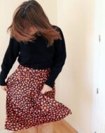 Mujer con jersey negro que, mirando hacia mueve la cabeza de un lado a otro haciendo que el pelo le tape la cara. Con sus manos está sujetando una falda fruncida, en color negro, con estampado de flores color coral rojizo, que recuerdan a las manchas de leopardo.