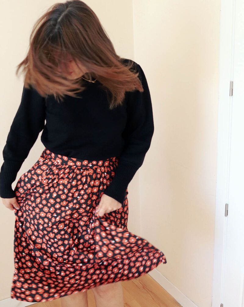 Mujer con jersey negro que, mirando hacia mueve la cabeza de un lado a otro haciendo que el pelo le tape la cara. Con sus manos está sujetando una falda fruncida, en color negro, con estampado de flores color coral rojizo, que recuerdan a las manchas de leopardo.