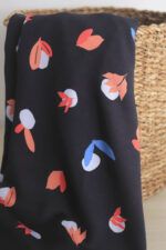 Guijarros en la cesta Una tela de color azul oscuro con motivos orgánicos de guijarros y hojas de diferentes formas en colores rojo, coral y azul, sobre sale y cuelga de una cesta redonda de esparto.