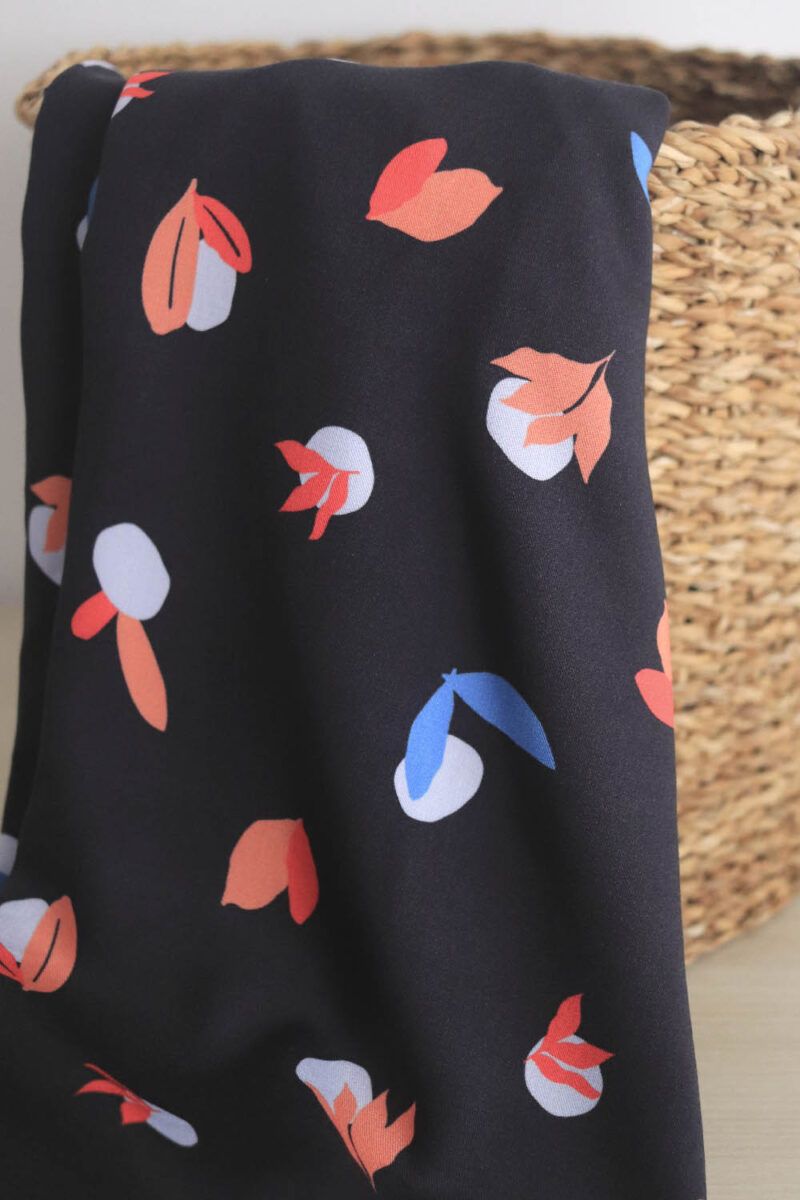 Una tela de color azul oscuro con motivos orgánicos de guijarros y hojas de diferentes formas en colores rojo, coral y azul, sobre sale y cuelga de una cesta redonda de esparto.