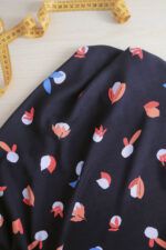 Guijarros metro Una tela de viscosa reposa sobre una mesa de madera de color claro. La tela es de color azul oscuro, casi negro, con motivos de guijarros en color azul muy claro, y diferentes tipos de hojas en colores coral, rojo y azul cian. A su lado se encuentra desenrrollada una cinta métrica de costura de color amarillo.