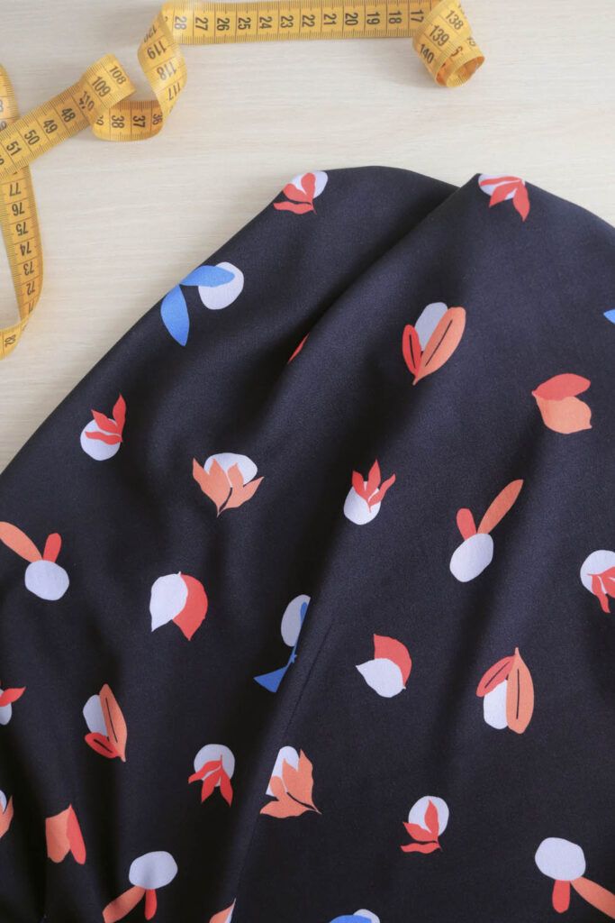 Una tela de viscosa reposa sobre una mesa de madera de color claro. La tela es de color azul oscuro, casi negro, con motivos de guijarros en color azul muy claro, y diferentes tipos de hojas en colores coral, rojo y azul cian. A su lado se encuentra desenrrollada una cinta métrica de costura de color amarillo.