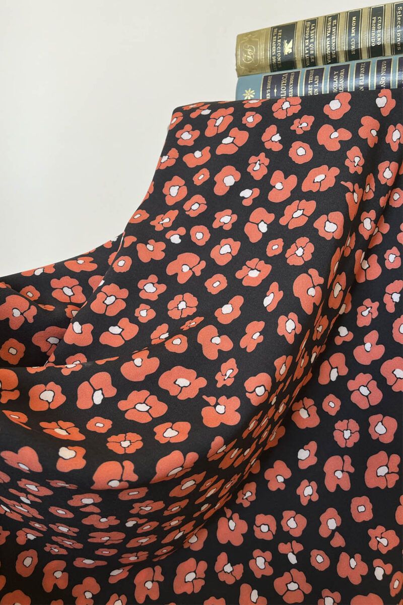 Una tela de fondo negro con flores que imitan las manchas de leopardo en color rojo coral y centro blanco cubre por completo un taburete. Sobre ella están, de manera horizontal dos libros antiguos de novelas en tonos verdes