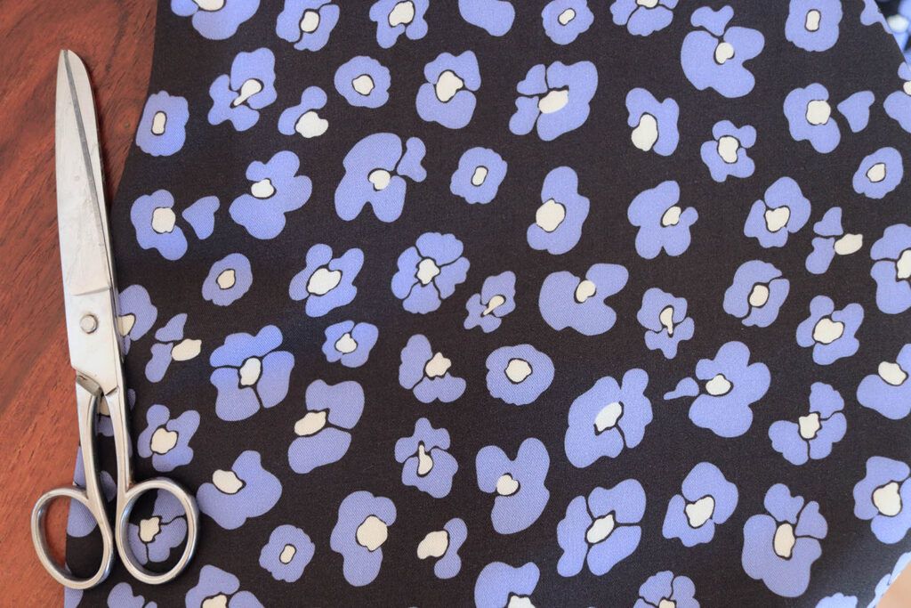 Tela de viscosa con fondo negro con flores que imitan las manchas de leopardo en color azul y centro color mantequilla sobre un fondo de madera oscura. Al lado hay una tijeras plateadas de costura