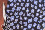 Tela con estampado de leopardo azul Tela de viscosa con fondo negro con flores que imitan las manchas de leopardo en color azul y centro color mantequilla sobre un fondo de madera oscura. Al lado hay una tijeras plateadas de costura