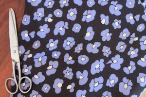 Tela de viscosa con fondo negro con flores que imitan las manchas de leopardo en color azul y centro color mantequilla sobre un fondo de madera oscura. Al lado hay una tijeras plateadas de costura