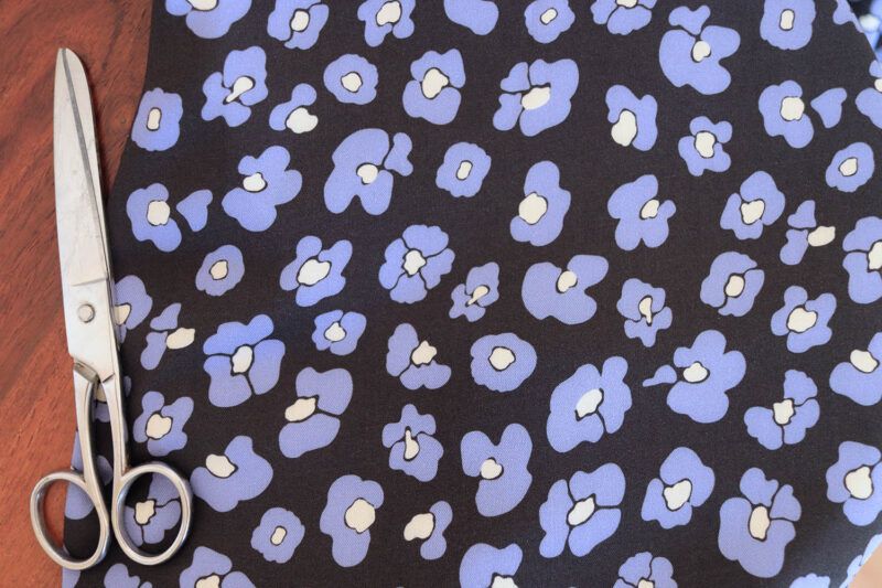 Tela de viscosa con fondo negro con flores que imitan las manchas de leopardo en color azul y centro color mantequilla sobre un fondo de madera oscura. Al lado hay una tijeras plateadas de costura