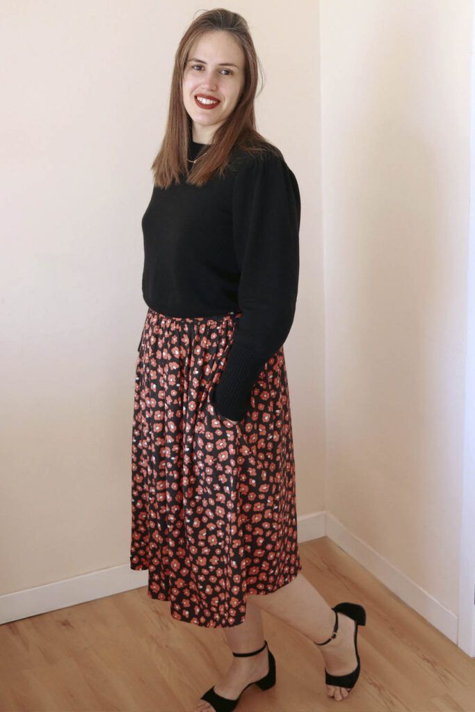 Mujer castaña, de pelo largo, posa sonriente ante la cámara. Está de medio perfil, lleva un jersey negro y una falda negra con estampado de leopardo en color coral. Sus manos están dentro de los bolsillos de la falda.