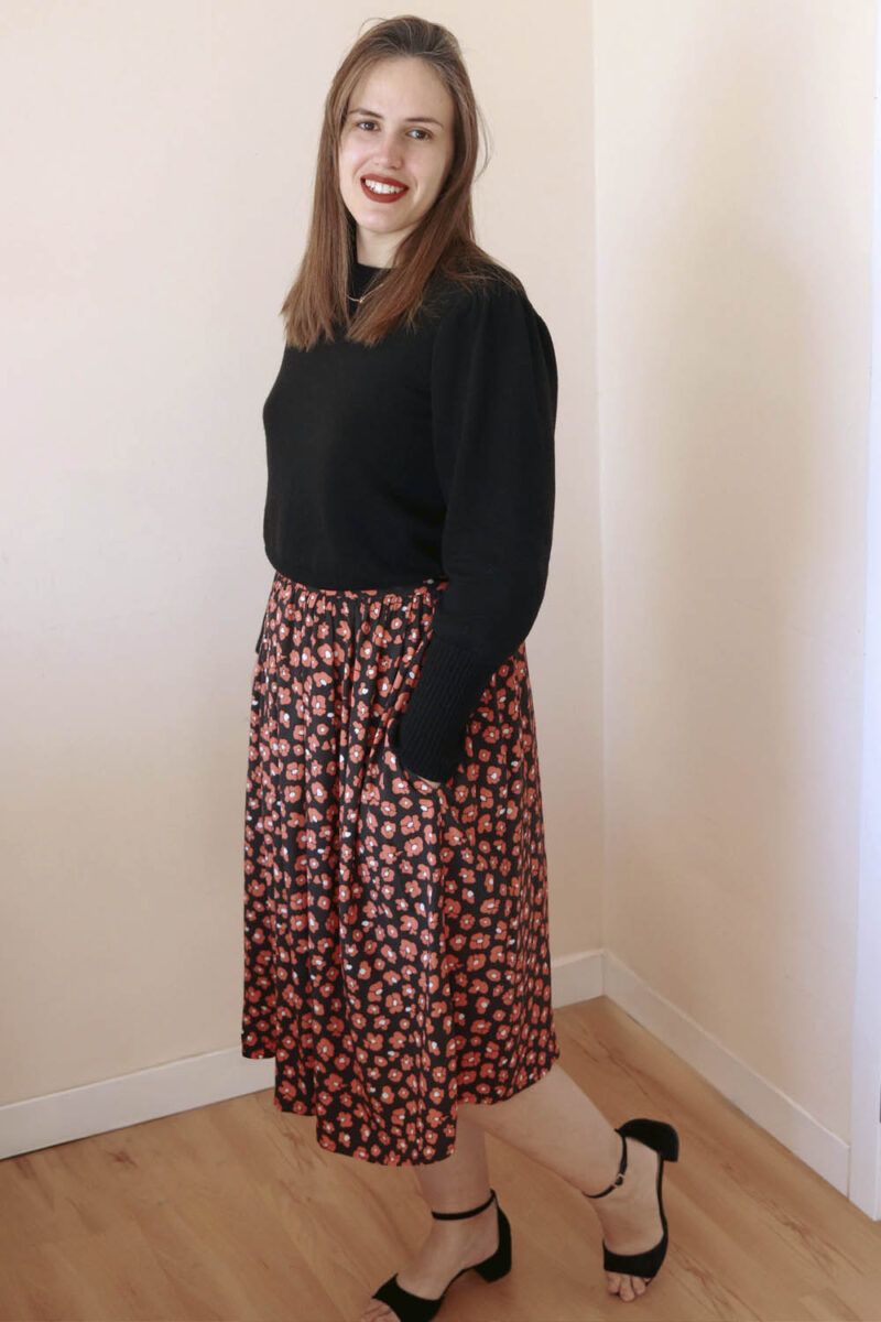 Mujer castaña, de pelo largo, posa sonriente ante la cámara. Está de medio perfil, lleva un jersey negro y una falda negra con estampado de leopardo en color coral. Sus manos están dentro de los bolsillos de la falda.