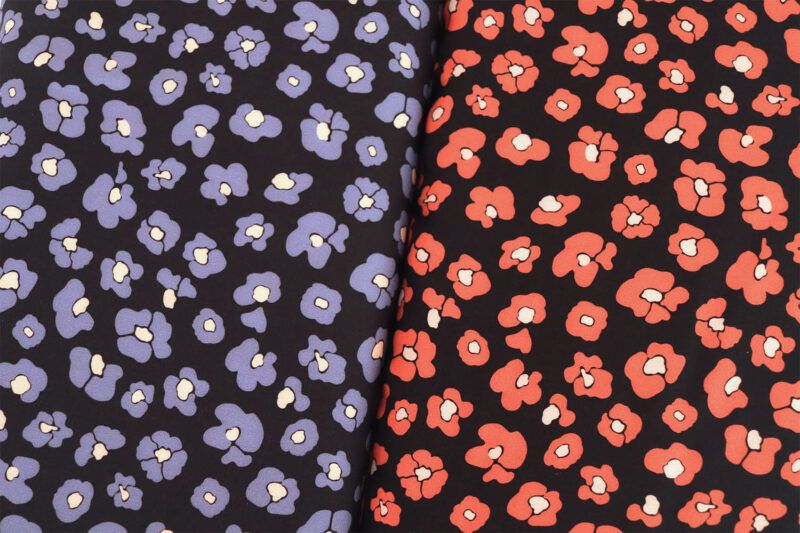 Dos tablillas de tela, una junto a la otra. Poseen el mismo estampado pero en diferentes colores. A la izquerda, una tela de fondo negro con flores que imitan las manchas de leopardo en color azul y centro color mantequilla muy claro. A la derecha, una tela de fondo negro con flores que imitan las manchas de leopardo en color rojo coral y centro blanco