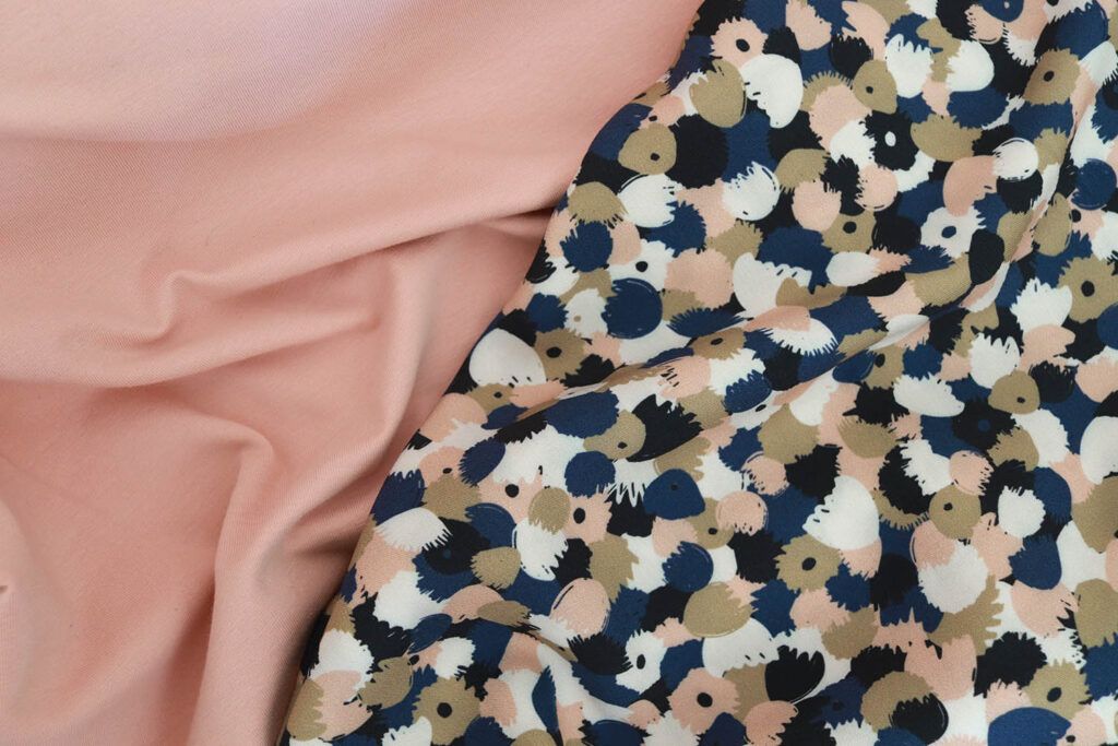 Primer plano de dos telas, una junto a la otra. A la izquierda se encuentra una tela de punto jersey de algodón en color rosa claro y a la derecha una viscosa con motivos de flores abstractas en colores azules, ocres, blancos y rosas. Ambas telas hacen diversos pliegues mostrando el comportamiento de la tela.