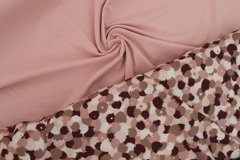 Primer plano de dos telas, una junto a la otra. A la izquierda se encuentra una tela de punto jersey de algodón orgánico en color rosa claro y a la derecha una viscosa con motivos de flores abstractas en colores burdeos, blancos y rosas. Ambas telas hacen diversos pliegues mostrando el comportamiento de la tela.