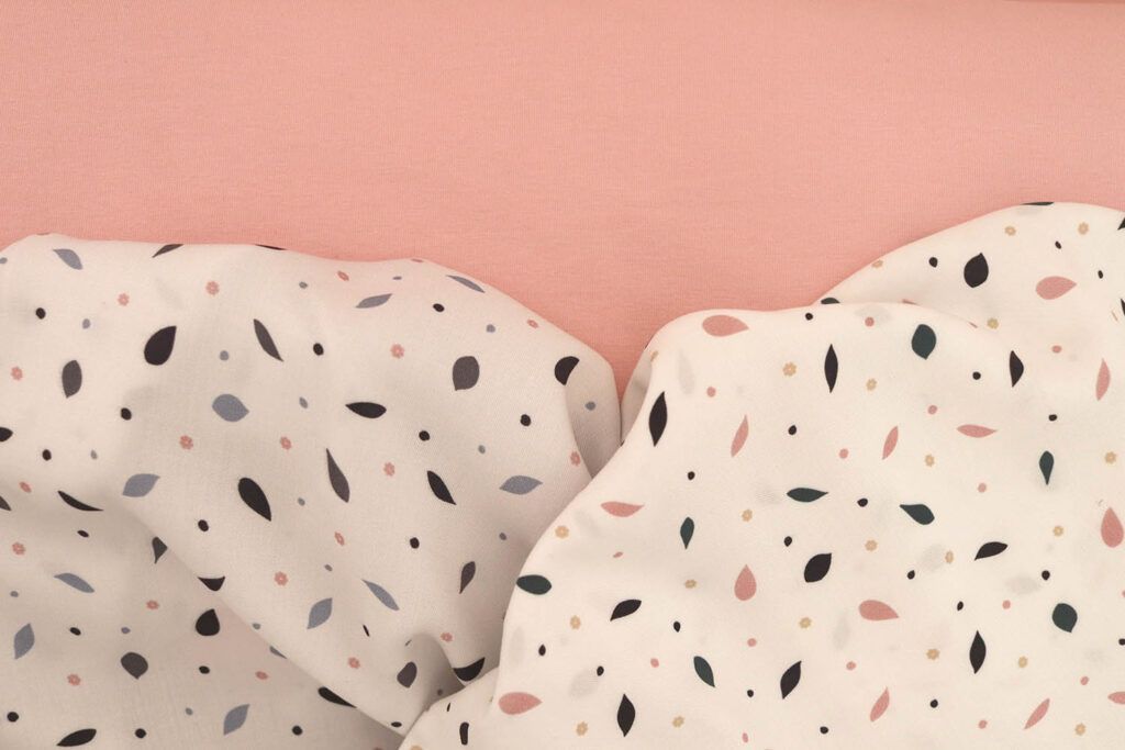 En la imagen se puede ver una plano cercano de tres telas juntas. Una es una tela de punto jersey de algodón en color rosa palo, otra es una viscosa de color blanco con motivos de hojas en distintos tonos azules y la otra es una viscosa con el mismo estampado que la anterior, pero las hojas son en tonos azules y rosas.