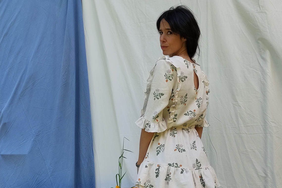 Elena OsesB, una mujer de cabello moreno, recogida, alta y estilizada posa frente a la cámara. Está de pie y de espaldas, pero gira su torso hacia la izquierda para mirar a la cámara. Lleva un vestido corto en color crudo con motivos de bouquets de flores en tonos verdes, naranjas y blancos. De fondo hay dos sábanas colgadas, a la izquierda una azul y a la derecha una blanca.